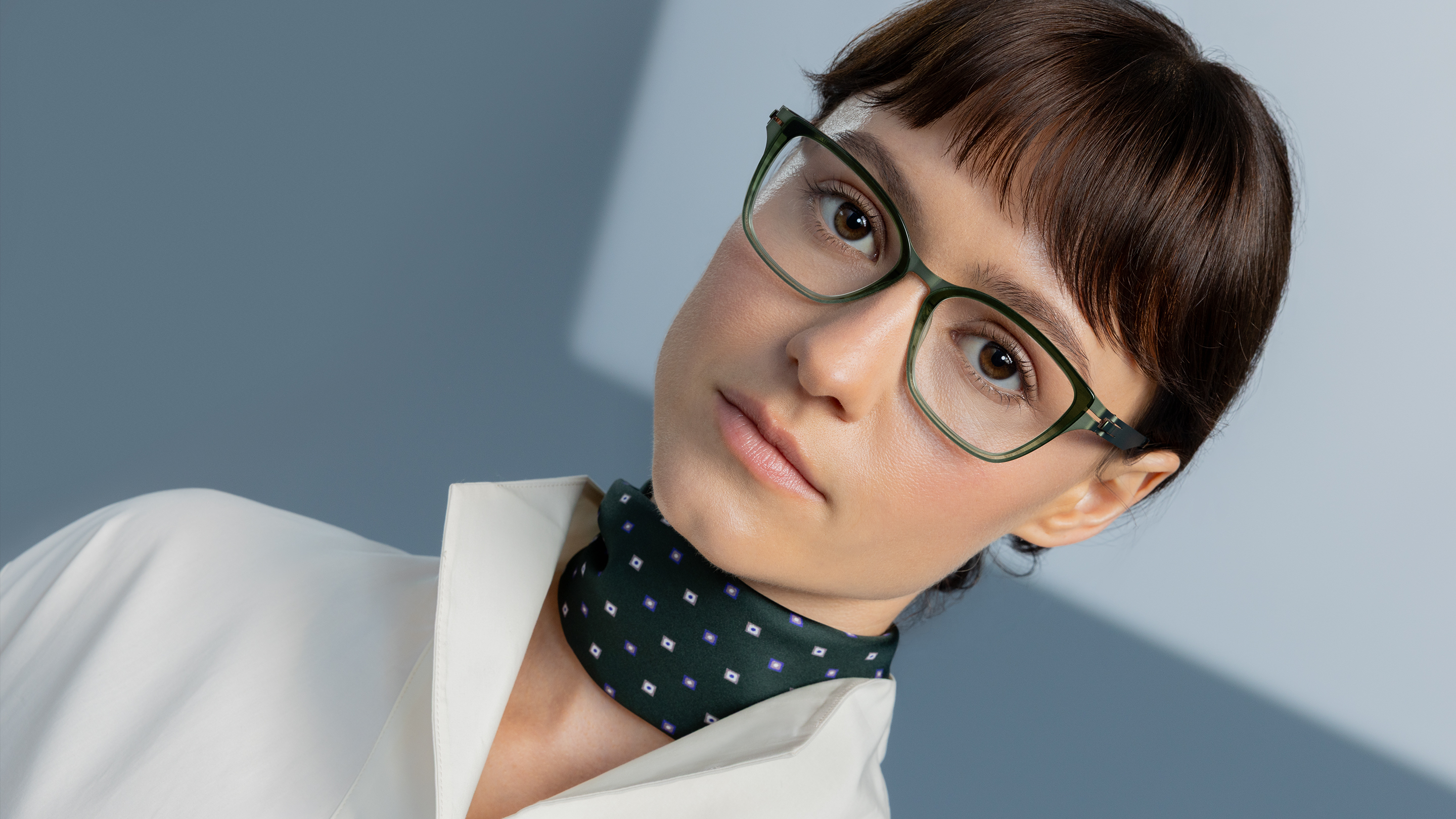 Woman in a white blouse and patterned green scarf wearing OVVO Optics DUO Marla translucent green eyeglasses against a blue-gray background